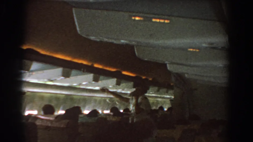 Wide 8mm frame of a 1967 aircraft cabin, showing seated passengers and an attendant reaching upward during the safety demonstration.