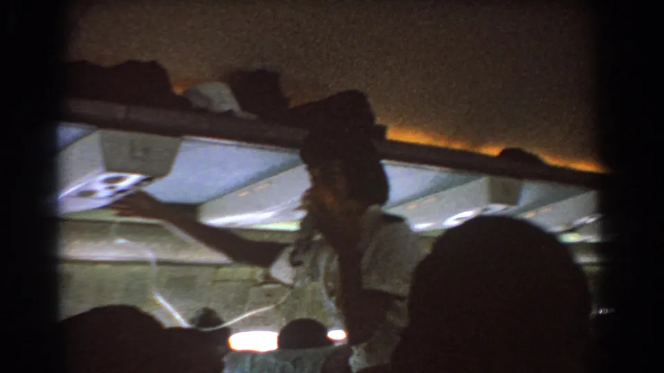 Closer cabin frame from 1967 showing a flight attendant demonstrating a dangling oxygen mask or line beside seated passengers.