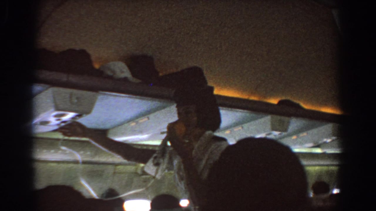 Flight attendant in an airplane cabin reaching toward overhead equipment as passengers sit nearby.