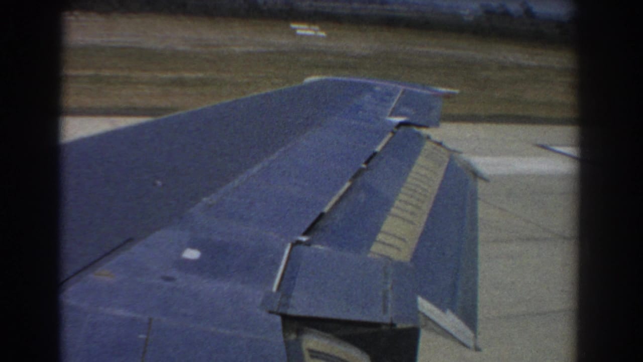 Blue airplane wing seen from a passenger window over runway and grassy airfield.