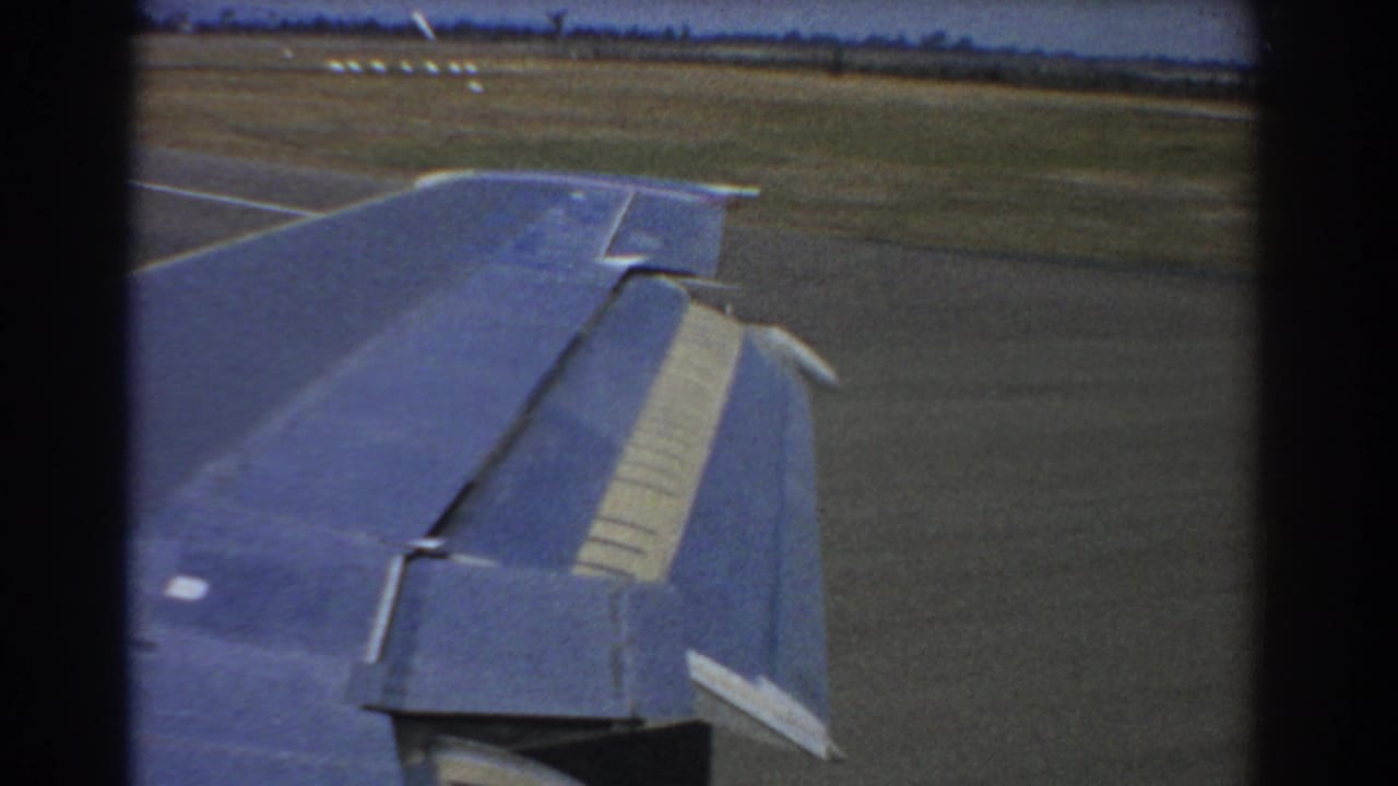 Blue airplane wing over runway pavement with grass and distant airfield visible beyond.
