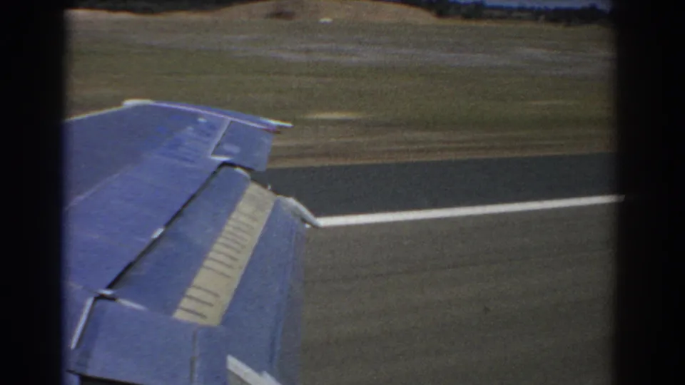 1967 8mm passenger view from a taxiing aircraft in Perth, showing the wing close to the window with a white runway line and blurred airfield grass.