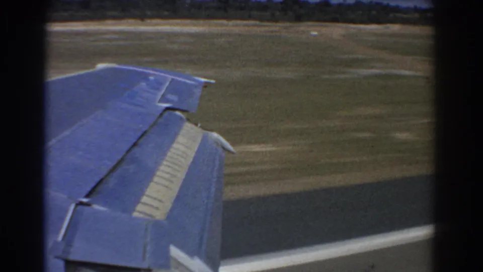 Late-1960s 8mm frame from Perth showing a blue airplane wing over dark runway pavement, with grass and airfield ground visible beyond the window.