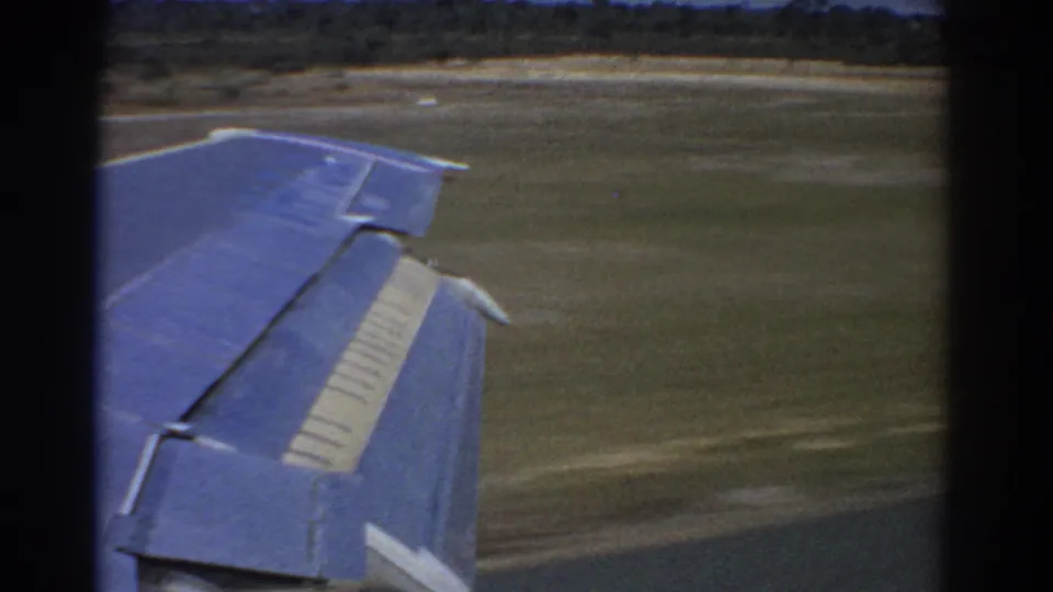 1967 Perth, Australia 8mm view from a passenger window showing an aircraft wing above grass near a runway.