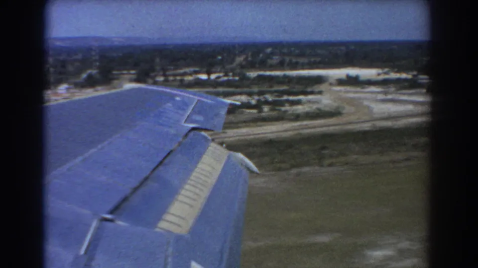 Passenger-window 8mm frame showing the aircraft wing above open grass, runway or service roads, and distant trees near Perth.