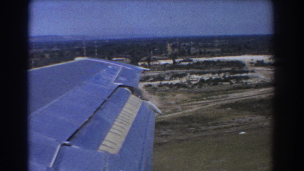 1967 Perth-area 8mm frame showing a blue aircraft wing in the foreground as the plane descends over roads, open ground and low buildings.