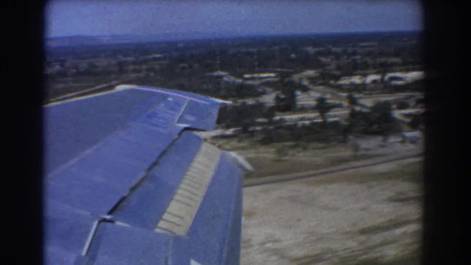 1967 Perth-area travel footage frame with a blue wing over a road network, scattered trees and low buildings as the aircraft nears the ground.