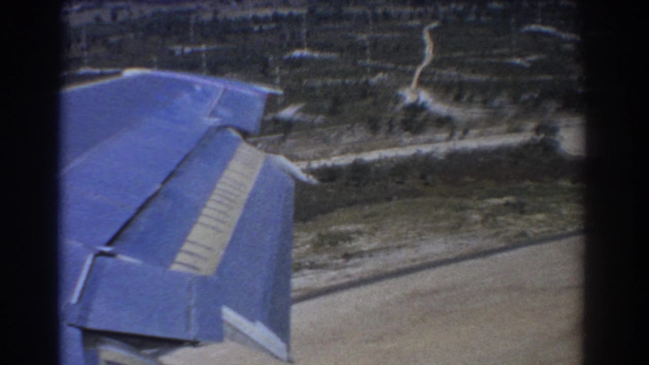Blue airplane wing seen over green farmland and pale dirt tracks.