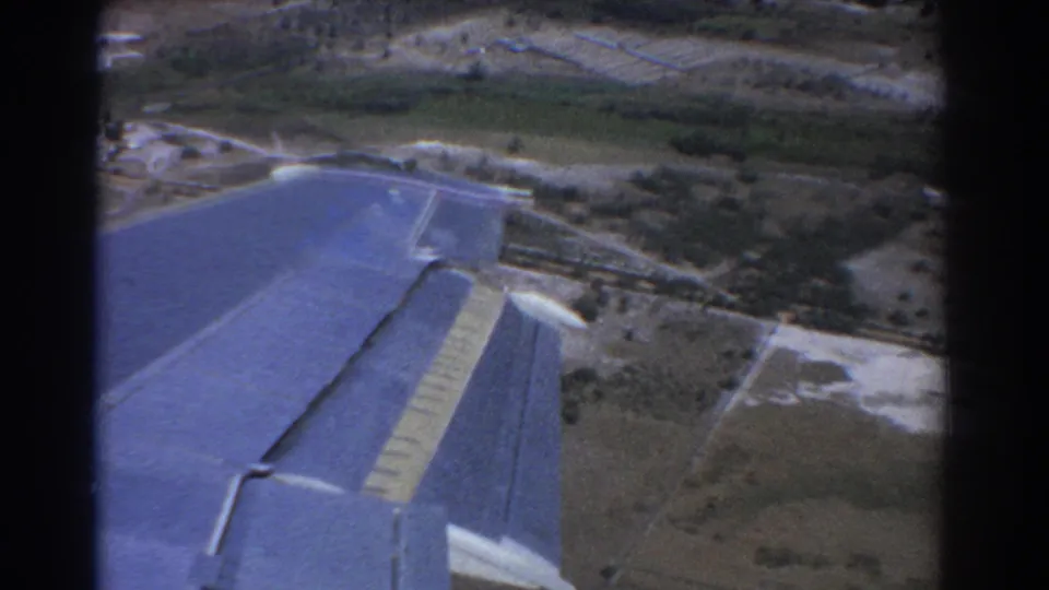 1967 aerial home-movie frame near Perth showing the blue aircraft wing over patchwork farmland, trees, and pale roadways.