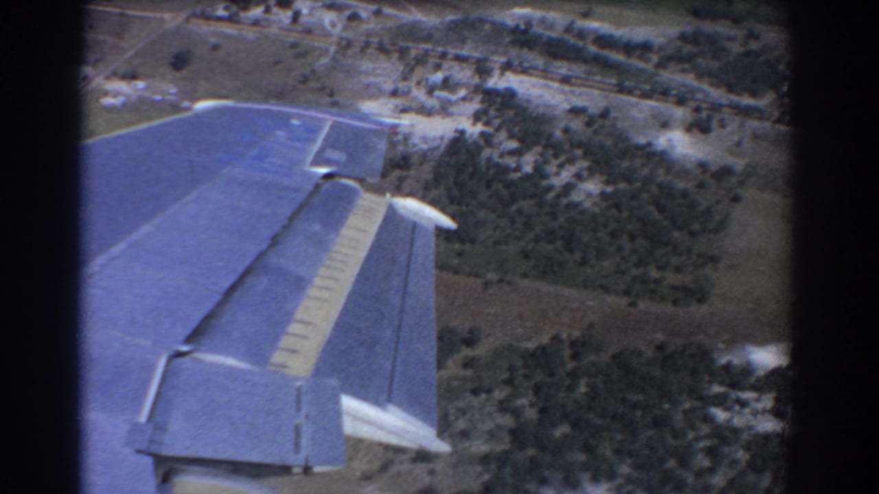 Aircraft wing above rural fields, trees, and pale ground tracks.