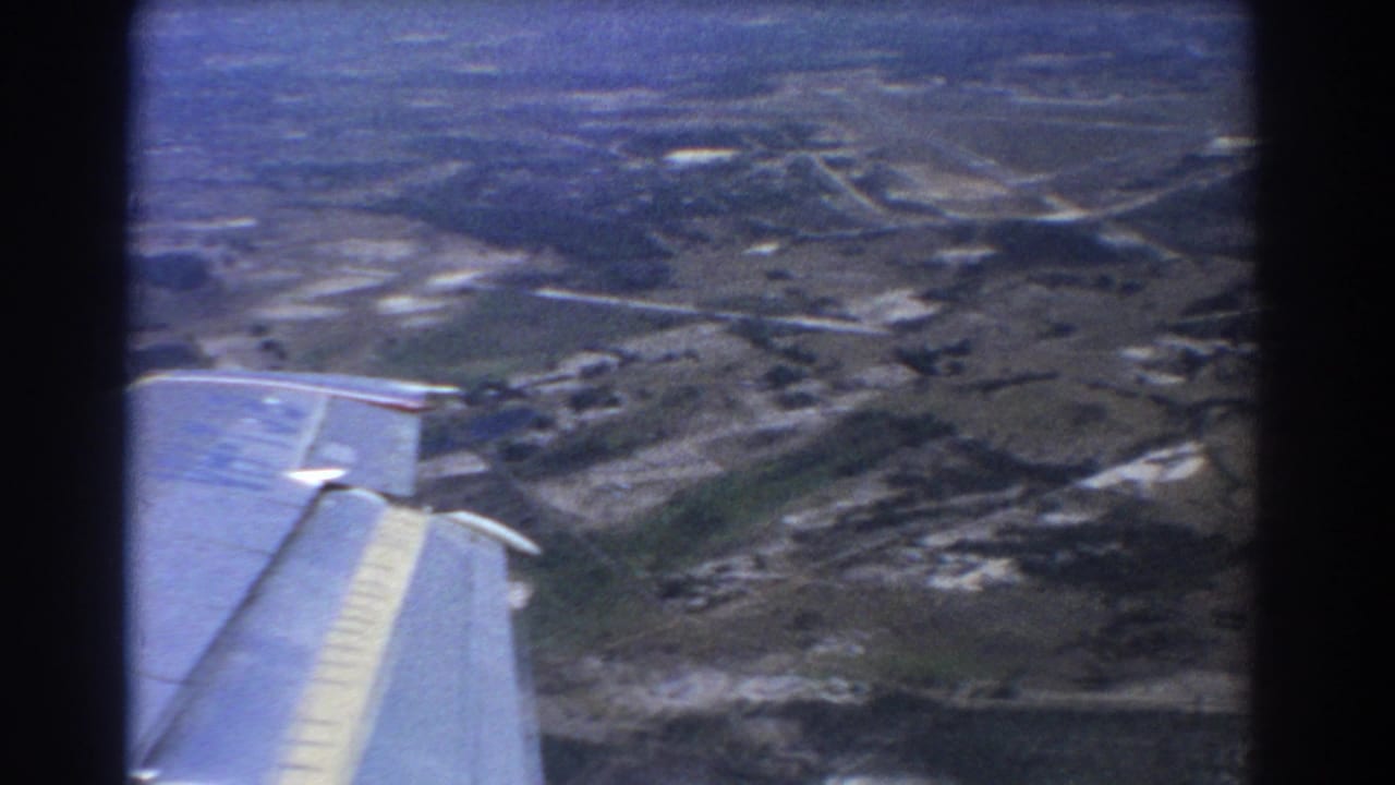 Airplane wing seen over fields and scattered structures from a passenger window.