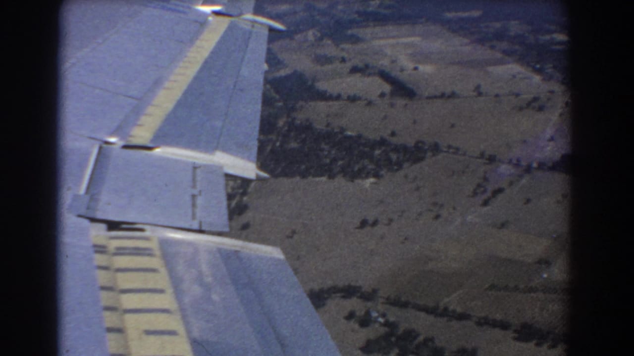 Aircraft wing and flaps over patchwork fields and tree lines.