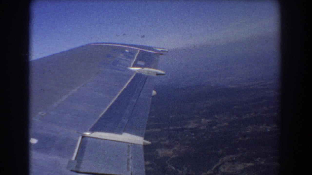 Airplane wing seen through a passenger window above hazy ground and blue sky.
