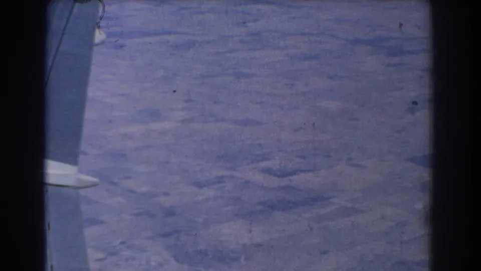 8mm passenger view looking past the aircraft wing and window edge toward pale patterned ground below.