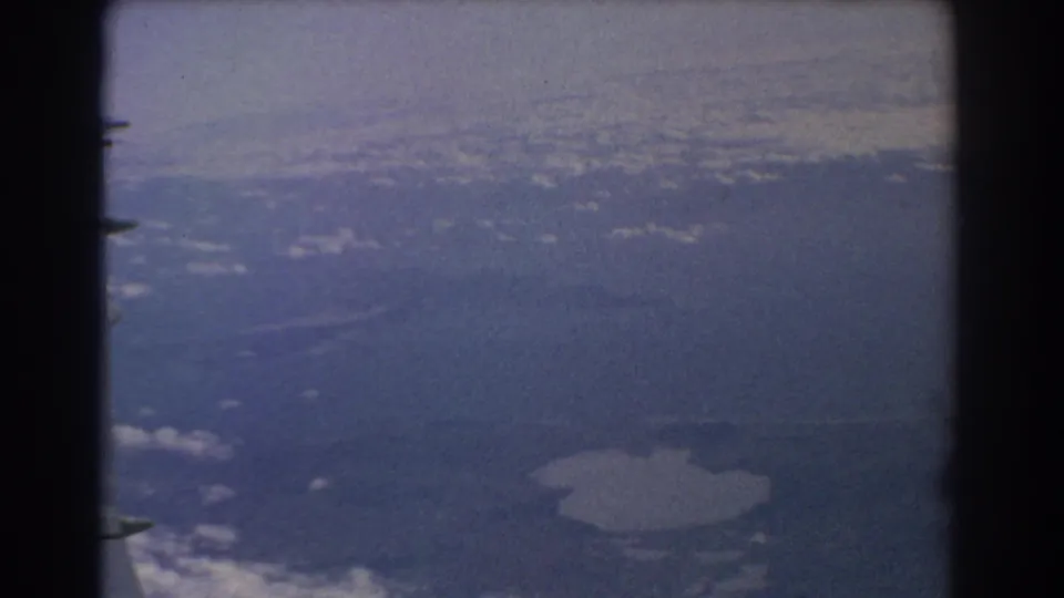 1967 8mm view from an airplane near Perth, showing a hazy landscape, scattered clouds, and lake water below.
