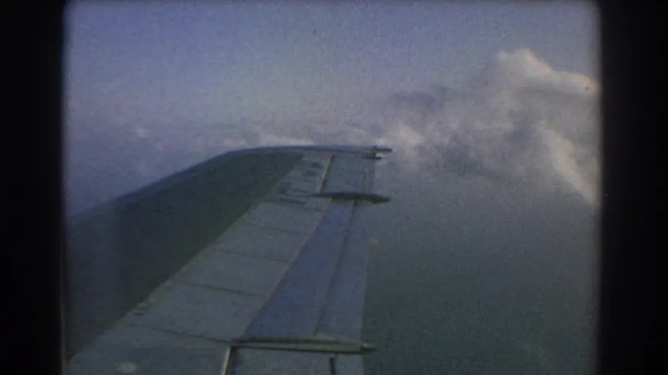 1967 8mm passenger view over Perth-area clouds, with the aircraft wing crossing a softly faded sky.