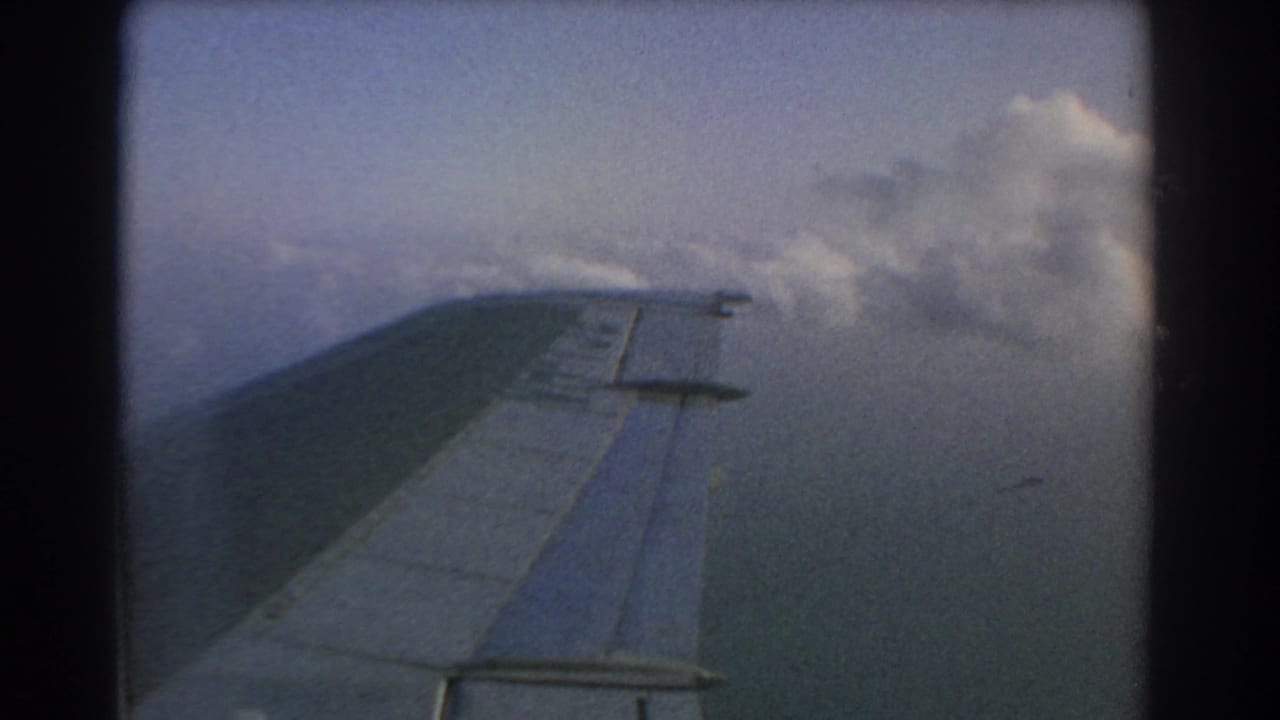 1967 8mm passenger-window view near Perth, Australia, showing an airplane wing over clouds and pale sky.
