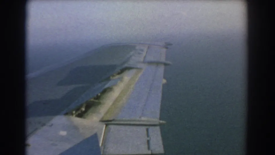 1967 8mm travel footage near Perth, Australia, showing an aircraft wing with extended slats above hazy blue water from a passenger-window viewpoint.