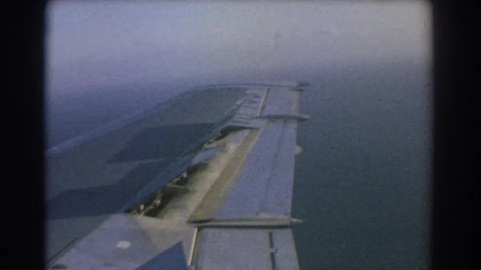 Passenger-window view from 1967 8mm footage showing the aircraft wing, raised slat surfaces, and muted blue water below during flight near Perth, Australia.