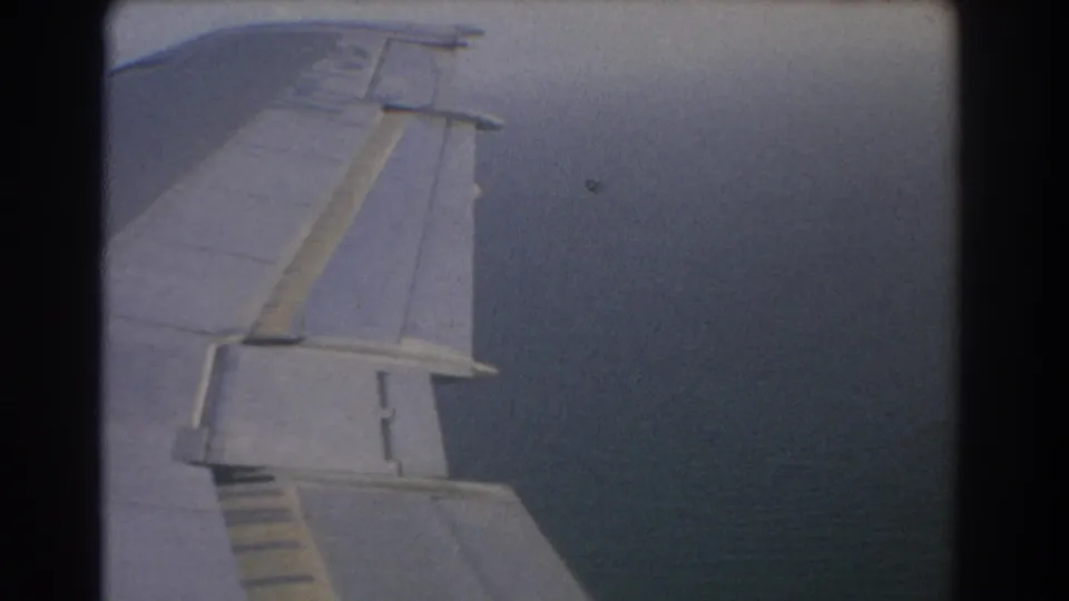 1967 8mm passenger view over Perth, Australia, showing an airplane wing and flap surfaces against a cloudy blue-gray sky.