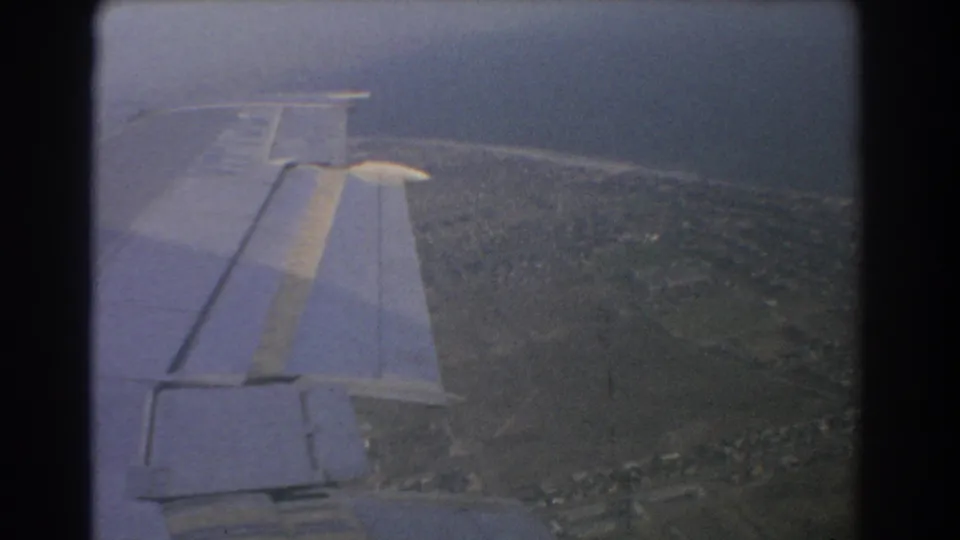 8mm airplane-window shot over Perth, showing a wing, built-up areas, roads, open land, and water in the distance.