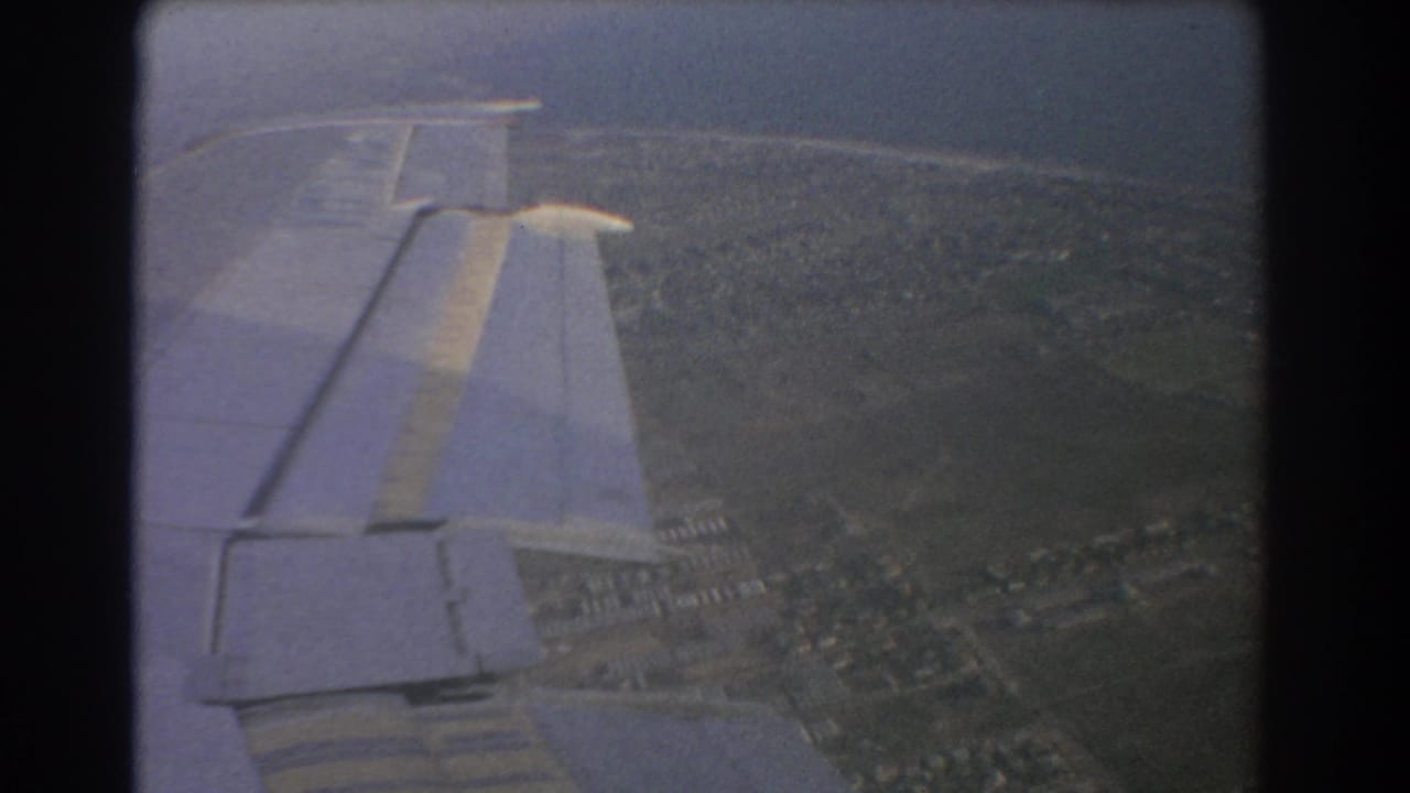 Airplane wing seen from a passenger window above green farmland and suburbs.