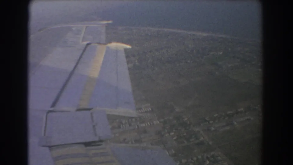 1967 Perth-area 8mm frame showing an airplane wing over green fields and faint suburban development below.