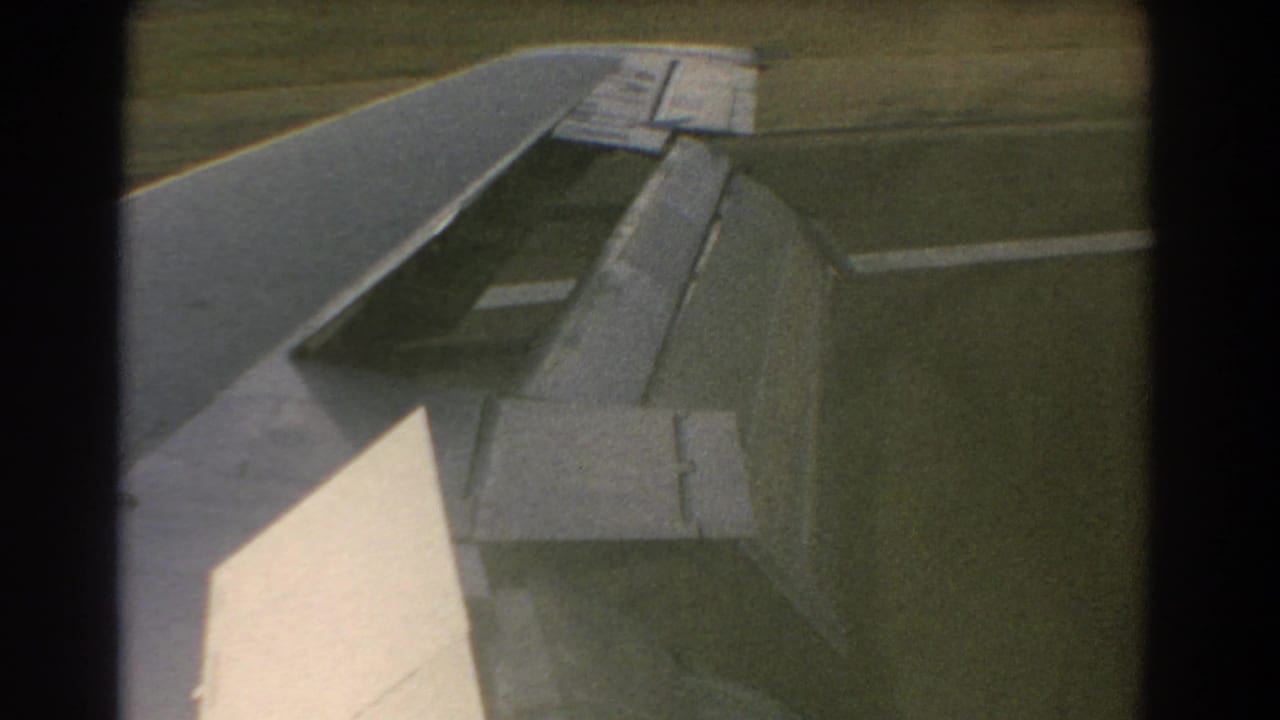 1967 Perth, Australia 8mm frame showing an airplane wing with flap extended above a grassy airfield.