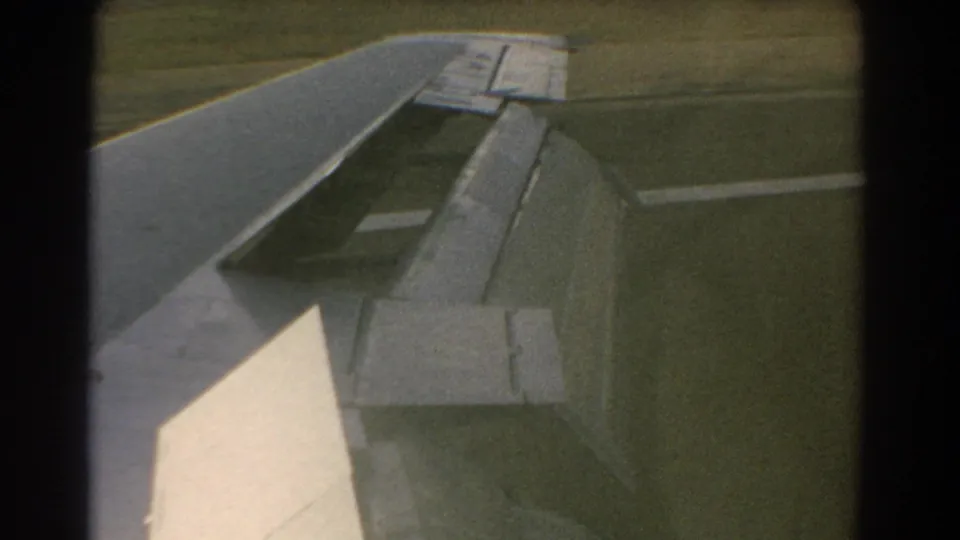 1967 Perth, Australia 8mm frame showing an airplane wing with flap extended above a grassy airfield.