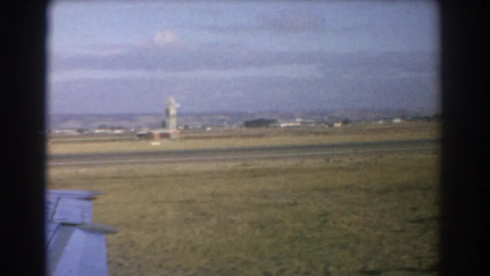 1967 Perth airport view with a distant control tower, low airfield buildings, grass, runway, and the aircraft wing edge.