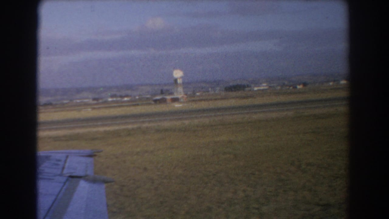 Aircraft wing over a grassy airfield with a distant dish-like tower and cloudy sky.