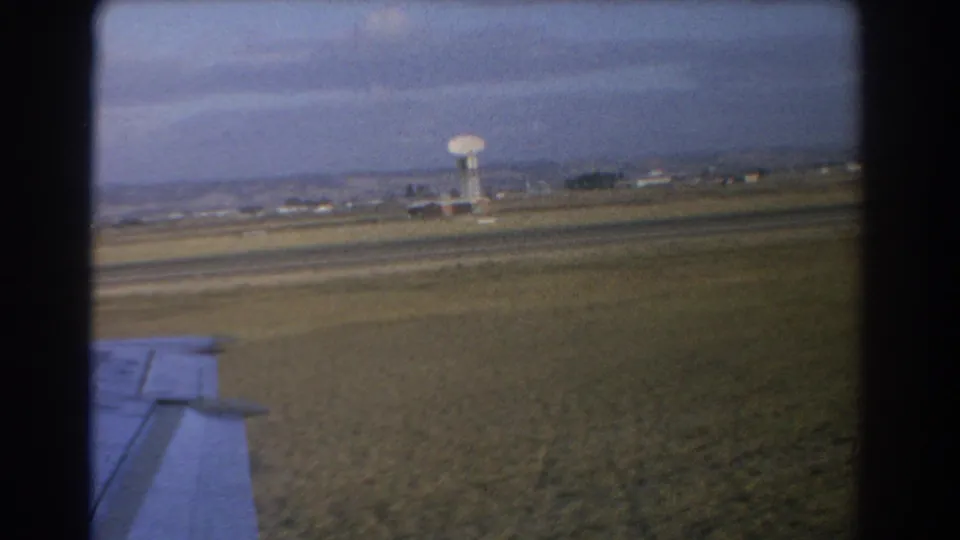 1967 Perth 8mm frame showing an airplane wing from the passenger window, flat airport grounds and a distant tower.