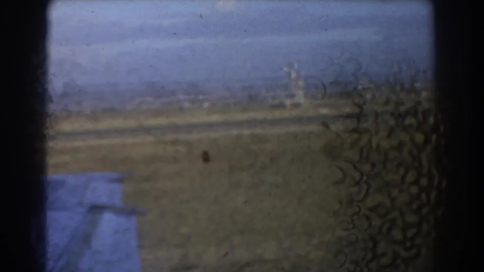 Soft 1967 Perth 8mm frame from an aircraft window, with the wing, runway ground and distant tower visible through film wear.
