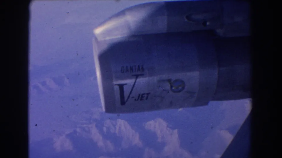 Passenger-view frame of a Qantas V-Jet engine and wing with hazy mountains visible through blue-tinted 8mm film.