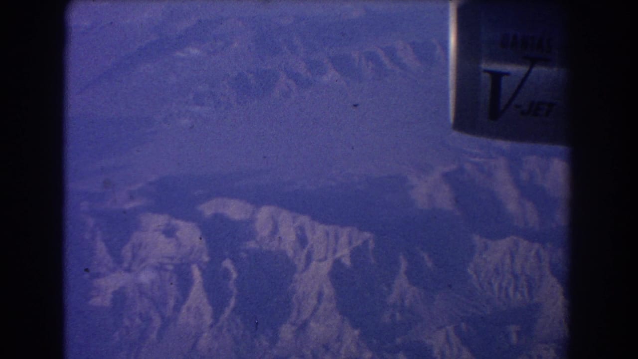 Blue-tinted aerial view of rugged mountains through an aircraft window.