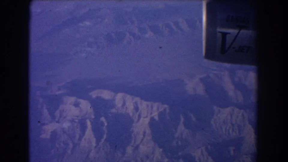 1967 8mm passenger-window view over rugged mountain ridges, with blue cast and part of the aircraft frame visible.