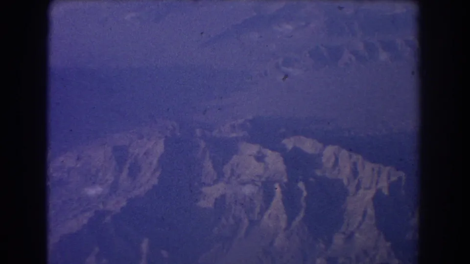 1967 home-movie frame showing hazy mountain terrain below, tinted blue by the film scan and window view.