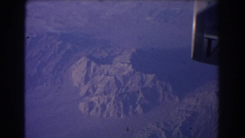 Blue 8mm frame of mountain ridges from a passenger aircraft, with partial Qantas V-Jet branding visible at the right edge.