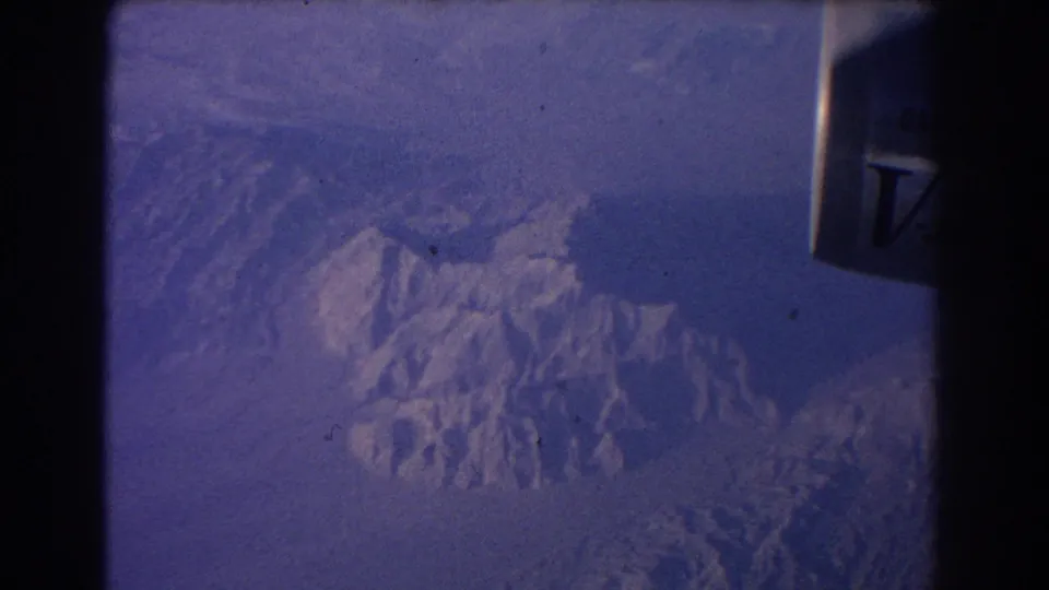 1967 8mm airplane-window view showing a jet engine edge over snow-dusted mountain peaks and hazy ridgelines.