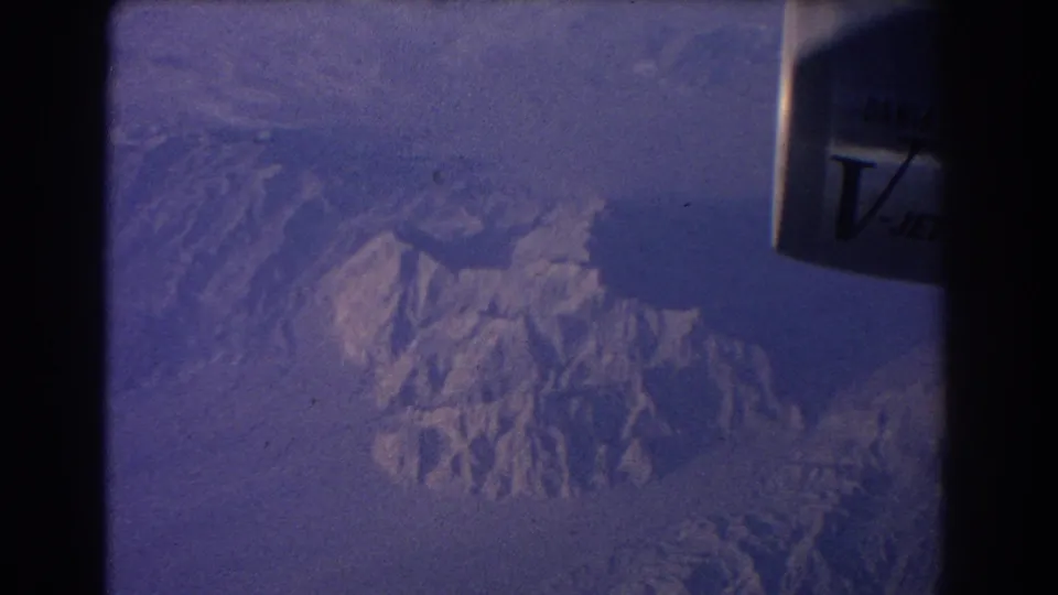 1967 8mm passenger view with dark window framing, partial jet engine lettering, and a rugged mountain range below.