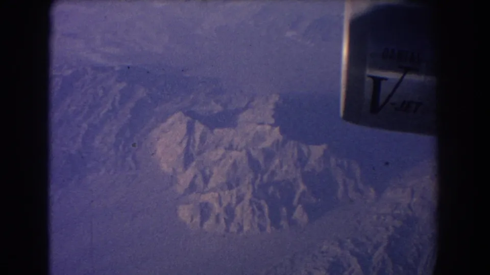1967 8mm view from a Qantas V-Jet window over snow-marked mountain ridges, with aircraft structure and frame edge visible.