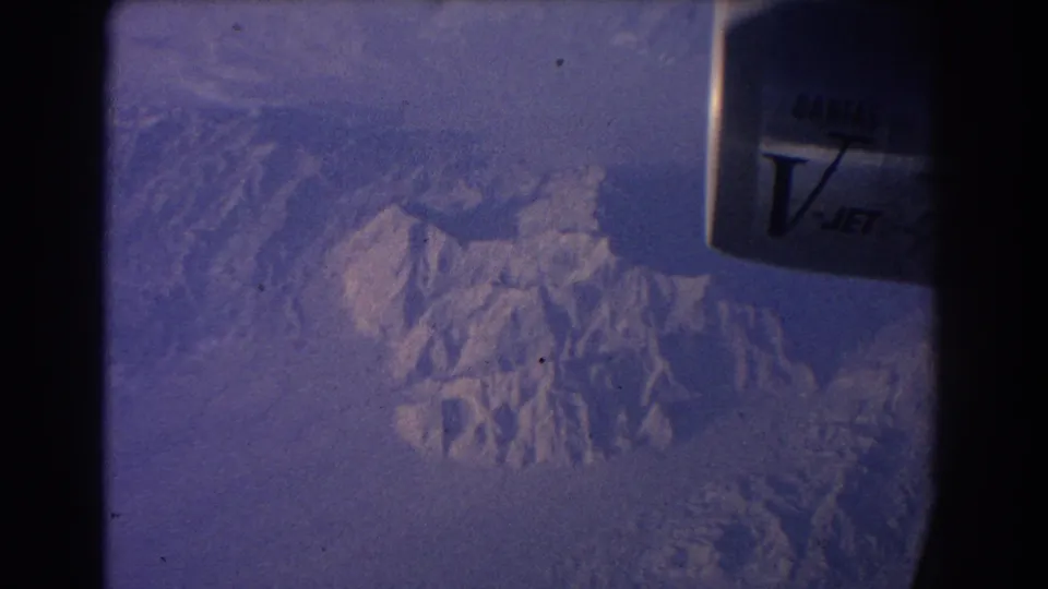 A steady 1967 passenger-window view shows rugged snow-capped mountains below and Qantas V-Jet lettering near the frame edge.