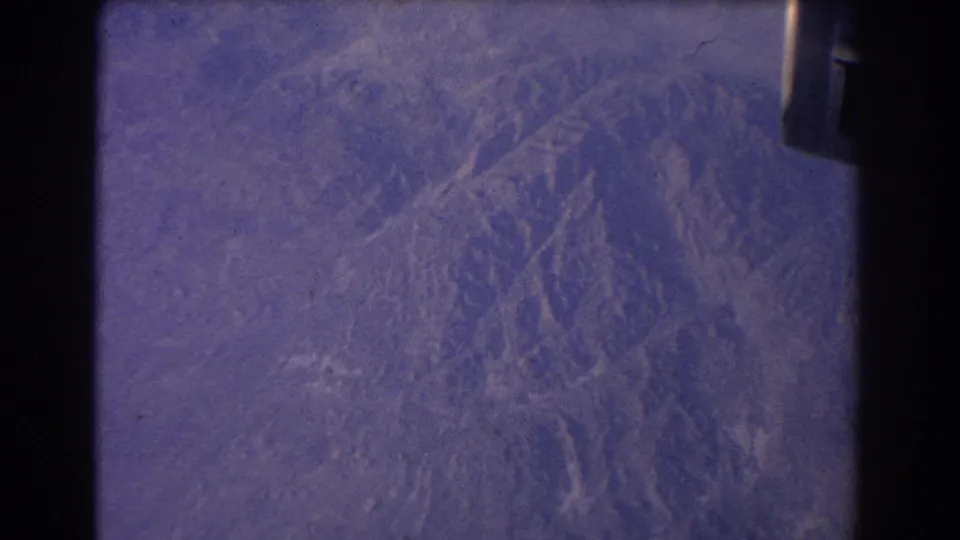 1967 8mm aerial view through an aircraft window, showing hazy ridges and valleys with a partial aircraft edge at right.