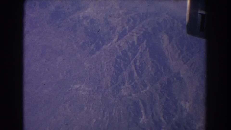 A 1967 8mm passenger view looks down on rugged mountain terrain, framed by a dark aircraft edge and faded blue-purple color.