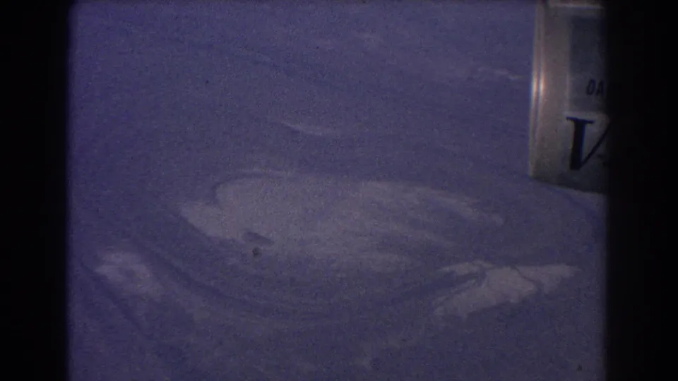 1967 8mm home-movie frame from the Sydney Australia clip showing ambiguous pale terrain or water patterns from above with a partial container edge.