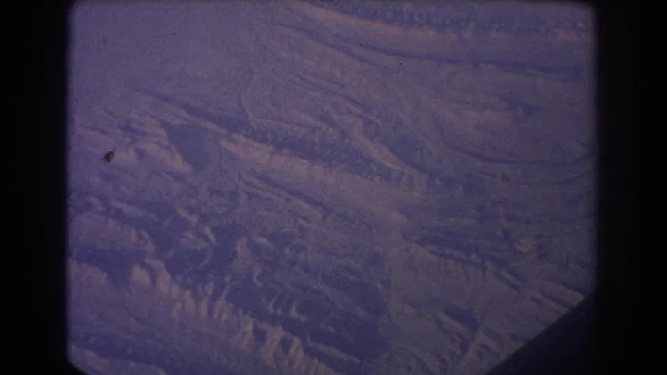 1967 8mm aerial view of snow-covered ridges and shadowed terrain, tinted purple-blue and framed by dark gate edges.