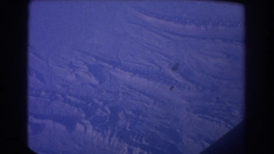 1967 8mm aerial view with bluish color shift, showing textured snowy terrain and shadowed ridgelines from above.
