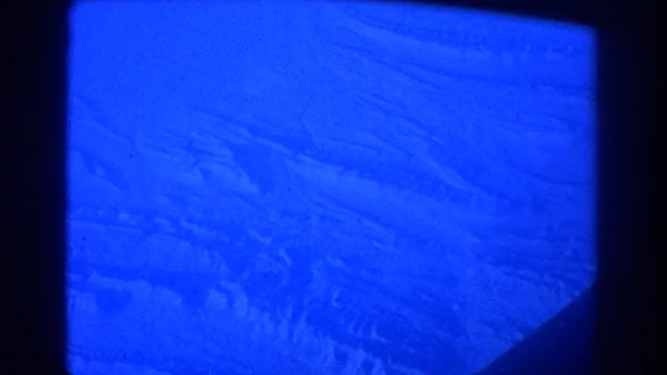 Deep blue-tinted 8mm frame of snow-covered uneven terrain viewed from above, with dark framing along the right and lower edge.