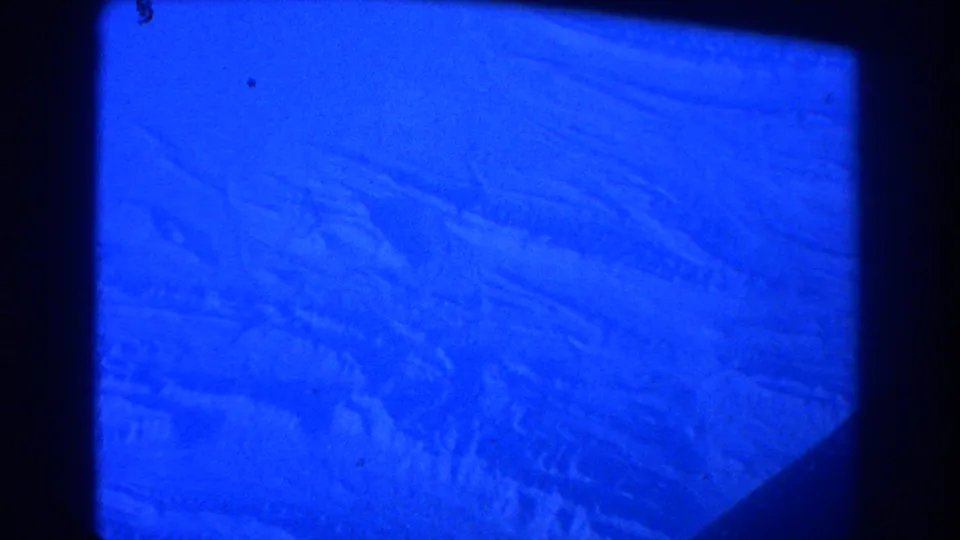 1967 8mm aerial view from an airplane window near Sydney, tinted deep blue with terrain and clouds faintly visible below.