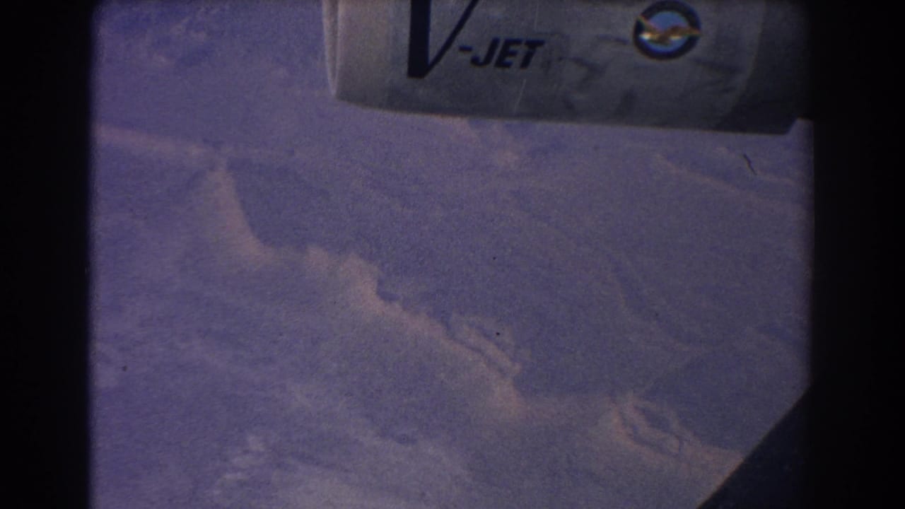 Aerial view of hazy mountain terrain through an aircraft window, with V-JET marking visible above.